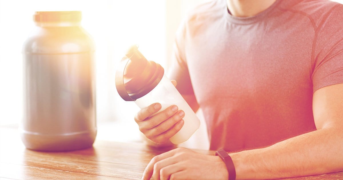 Man holding up a protein shaker with protein container in the background.
