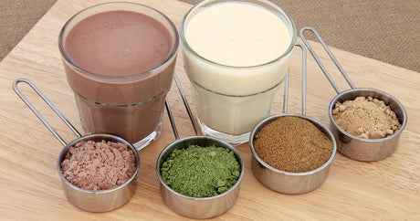Two glasses filled with white and brown content as well as 4 cups of different plant protein powder.