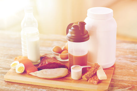 Wooden cutting board with different protein sources like eggs, salmon, chicken breasts, milk and protein powder on top