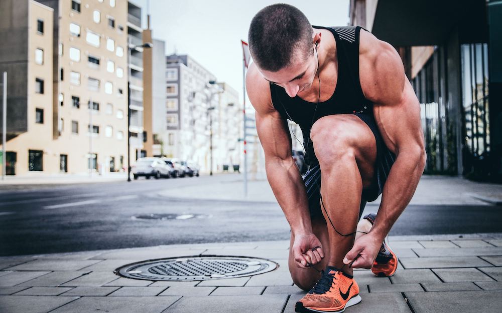 A man exercising in the city, knelt on the footpath tying his sneaker shoelace