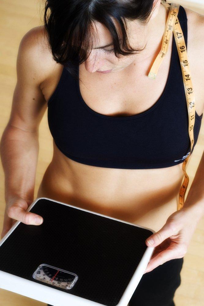 Woman in a sports bra with a measuring tap on her shoulder are holding a scale