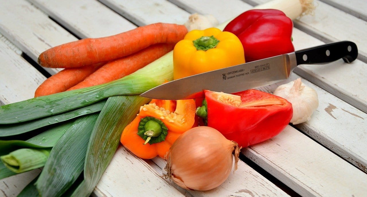 Carrots, different colours capsicums, an onion and leek as well as a big sharp knife are lying on a wooden surface